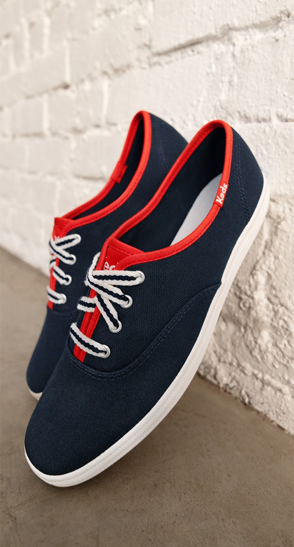 A pair of Keds Champion Canvas Varsity Lace shoes propped up on a white brick wall